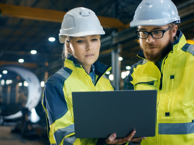 Two people in hard hats looking at a laptop in a factory.