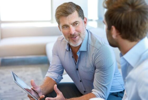 Two men in business attire sitting as they discuss something. One of the men holding a tablet in his right hand.