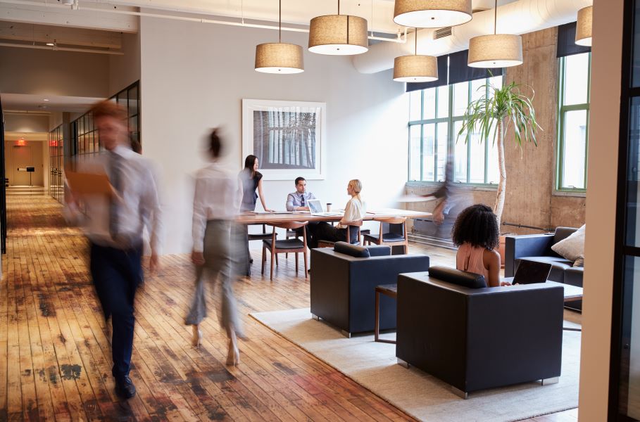 A modern office space with a wooden floor, some people seen clearly as they sit at desks or in leather chairs, others blurred as they walk across the floor.