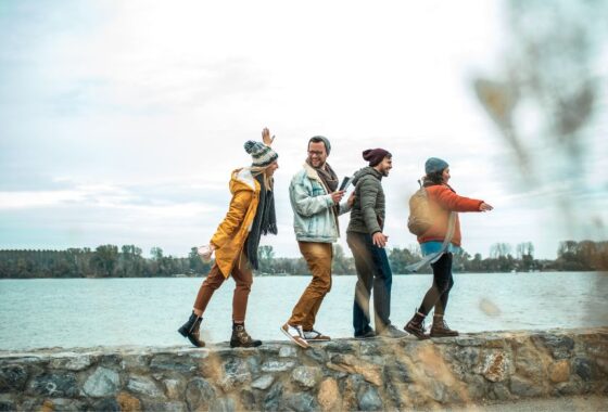 Four hikers walking playfully along a low wall in front of a body of water.