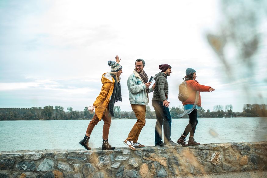 Four hikers walking playfully along a low wall in front of a body of water.