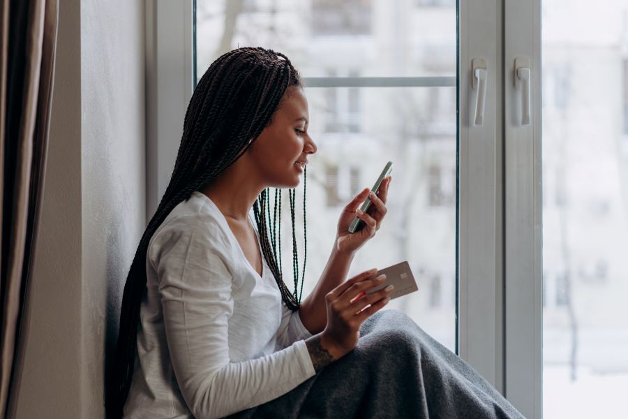 A woman sat by a balcony door, a phone in each hand, as she looks down at the phone in her left hand.