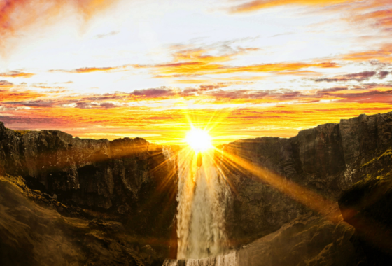 The sun, bright but low in the sky, just above a waterfall that is the centrepiece of a rocky landscape.