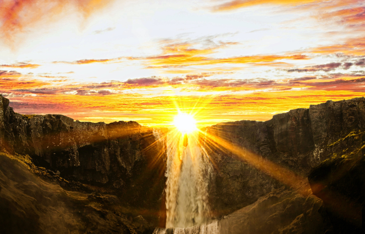 The sun, bright but low in the sky, just above a waterfall that is the centrepiece of a rocky landscape.