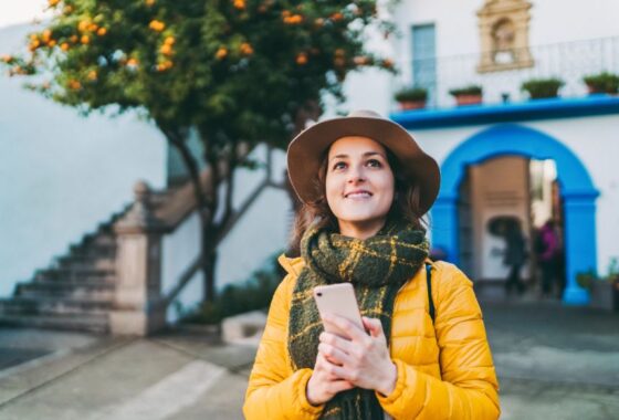 A woman in a yellow hat and scarf is looking at her phone in front of a building.