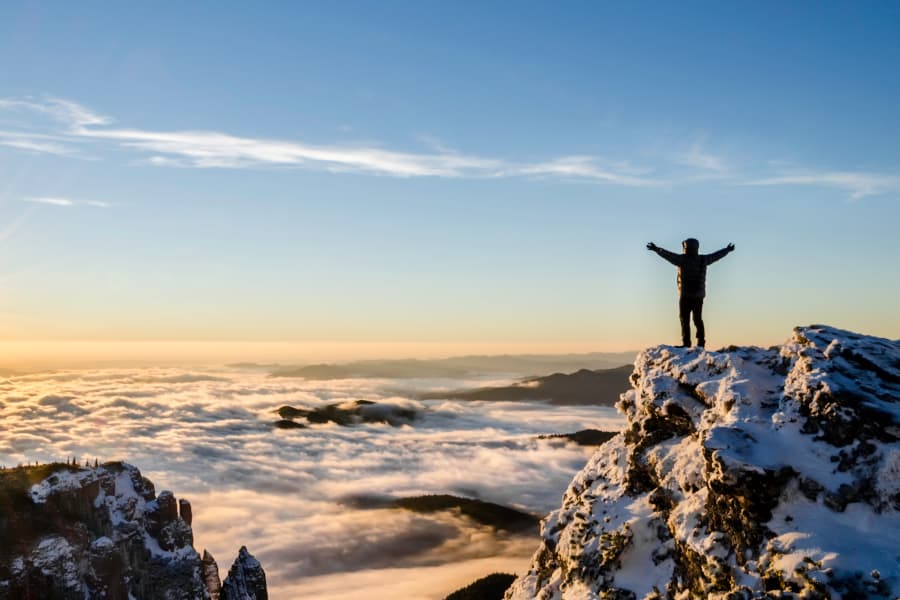 A person standing on a high, snow-covered rocky outcrop, arms outstretched, clouds or mist obscuring much of the landscape below them, with the occasional peak rising through.