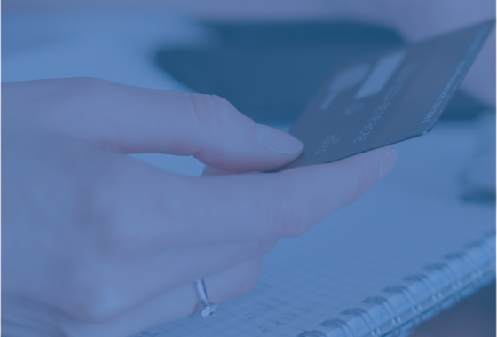 A close-up of a hand holding a credit card, as if examining, or entering the details on a PC. The whole image has a blue hue.