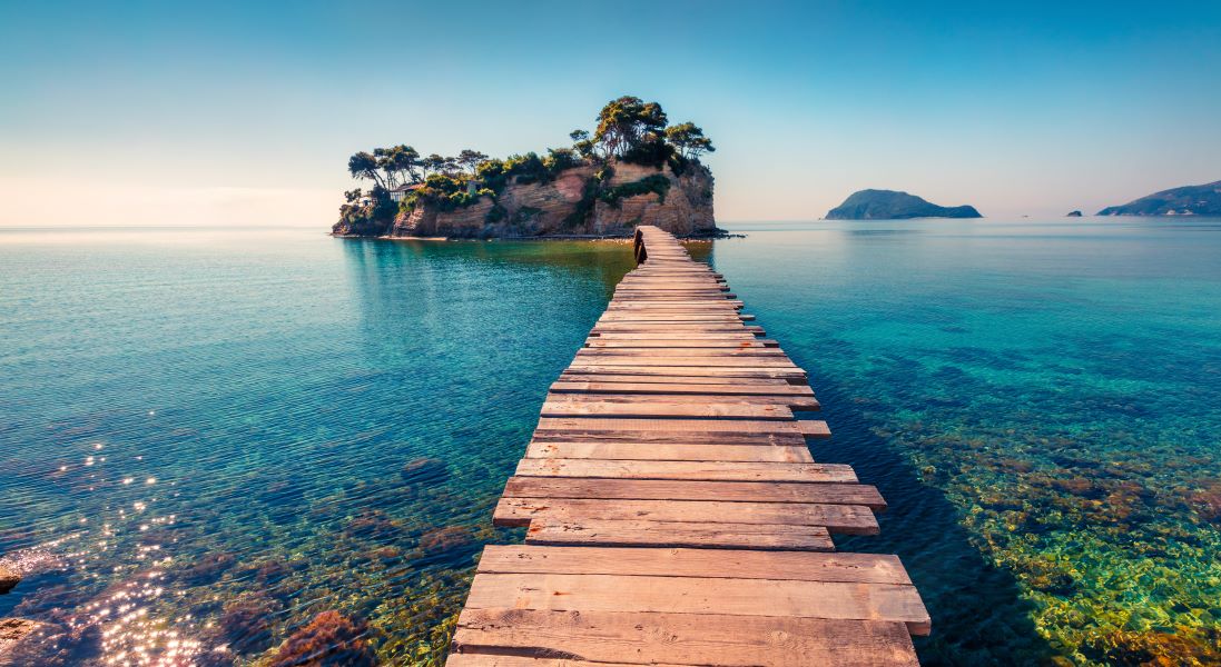 A rustic wooden boardwalk stretches away from us and over bright blue water towards a rocky island.