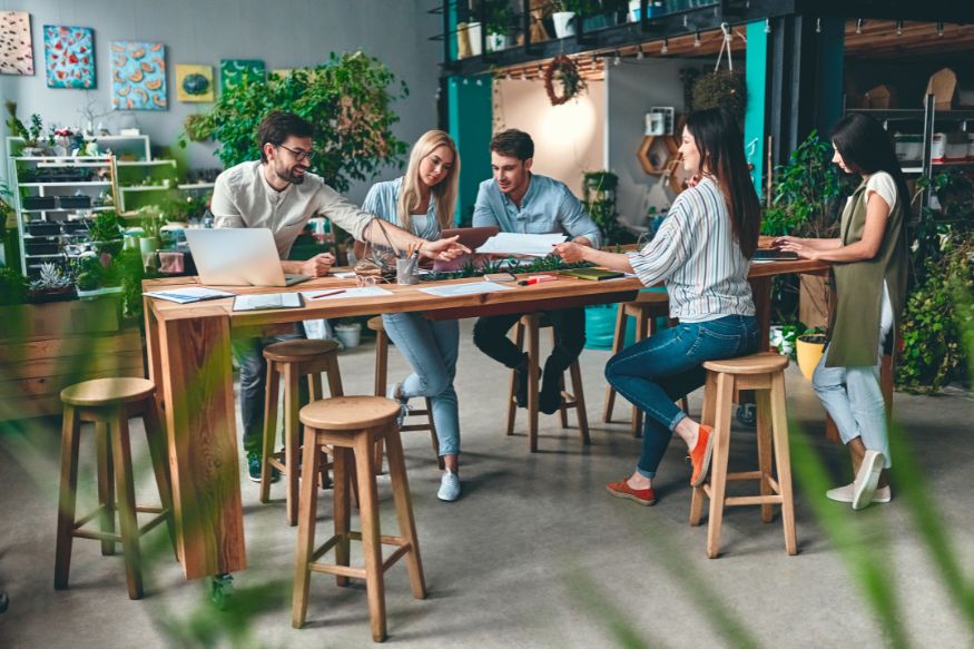 A group of 5 people gathered around a desk with numerous pieces of paper, items of stationery and a laptop on it, as if working together.