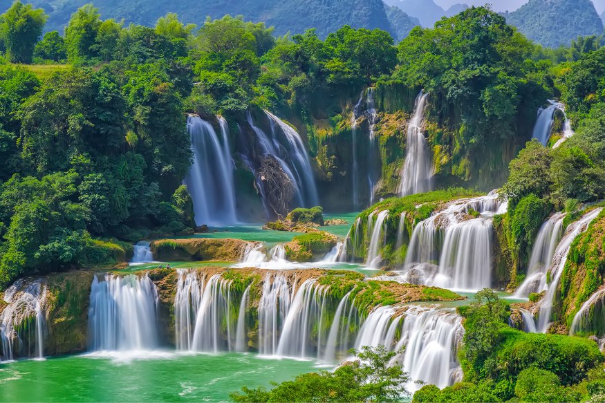 A three-tiered waterfall in a rainforest, turquoise water flowing down and between rocks and foliage into a large body of water at the bottom.