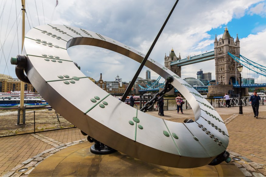 A large, metal, sundial style clock with London's Tower Bridge in the background.