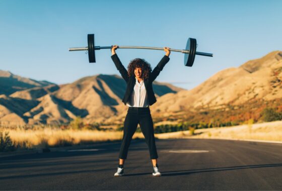 A woman in a business suit lifting a barbell in the middle of the road.