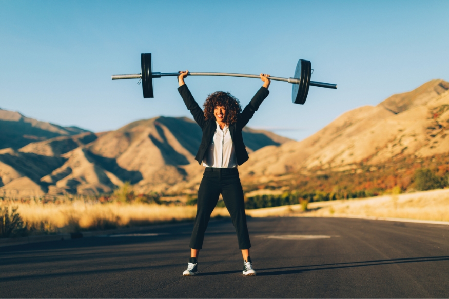 A woman in a business suit lifting a barbell in the middle of the road.