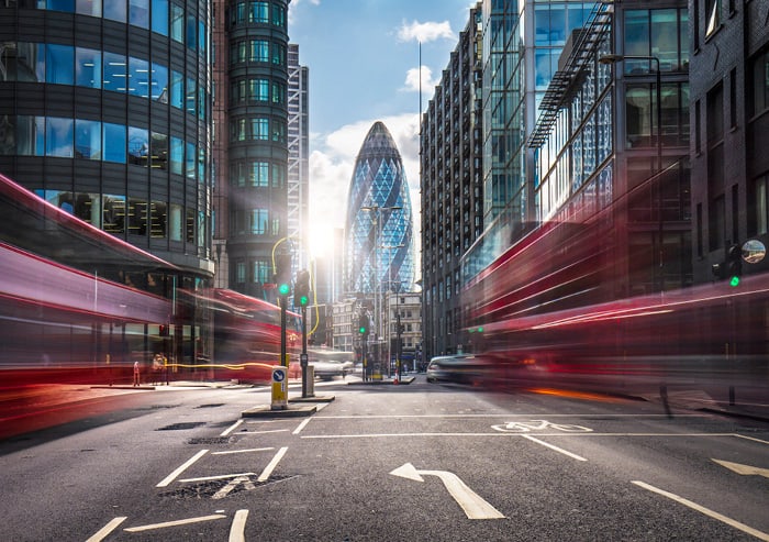 A busy Central London street in the daytime, buses heavily blurred as they pass on either side, 'The Gherkin' visible in the centre of shot in the distance.