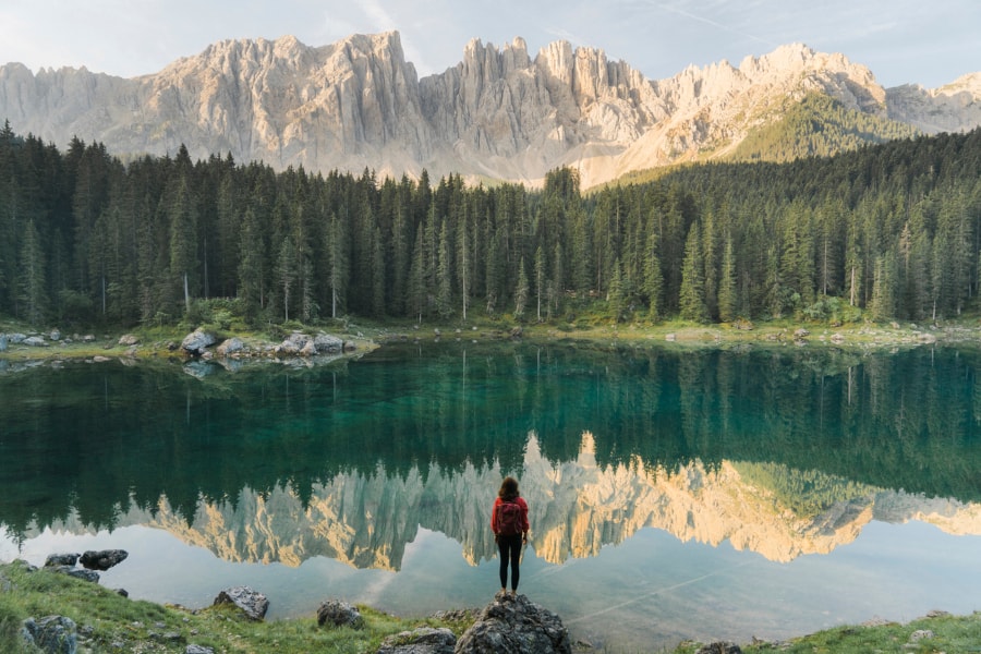 A female hiker standing on a rock looking at a still lake, a dense forest on the other side of the lake, and mountains further in the distance.