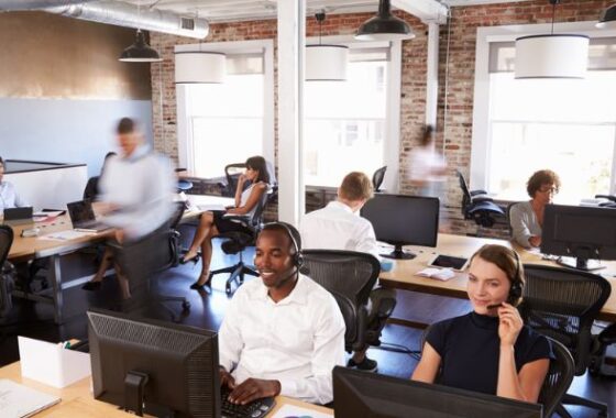 A busy office scene, two people wearing headsets in the foreground, further colleagues behind them, most sat at desks using computers, but some are blurred as they walk.