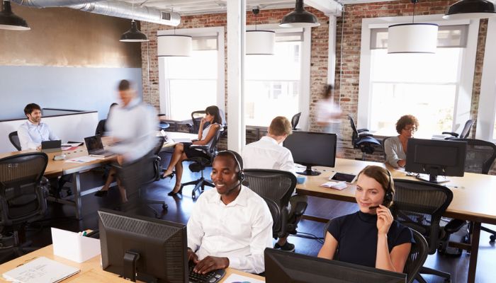 A busy office scene, two people wearing headsets in the foreground, further colleagues behind them, most sat at desks using computers, but some are blurred as they walk.