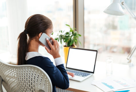 A woman seen from behind, sat at a desk working on a laptop and holding a phone to her ear.