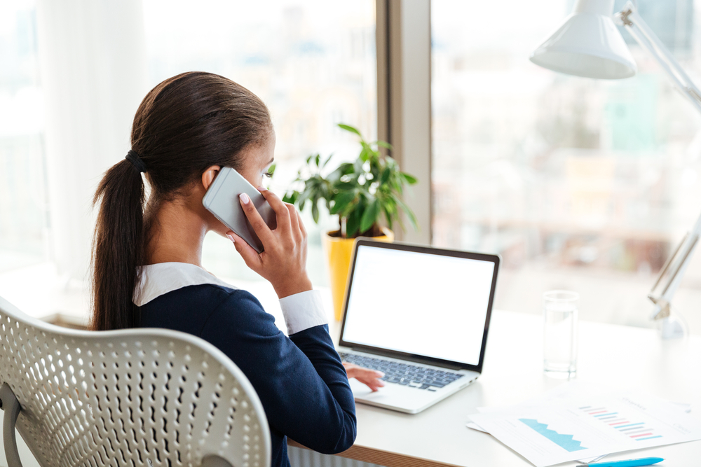 A woman seen from behind, sat at a desk working on a laptop and holding a phone to her ear.