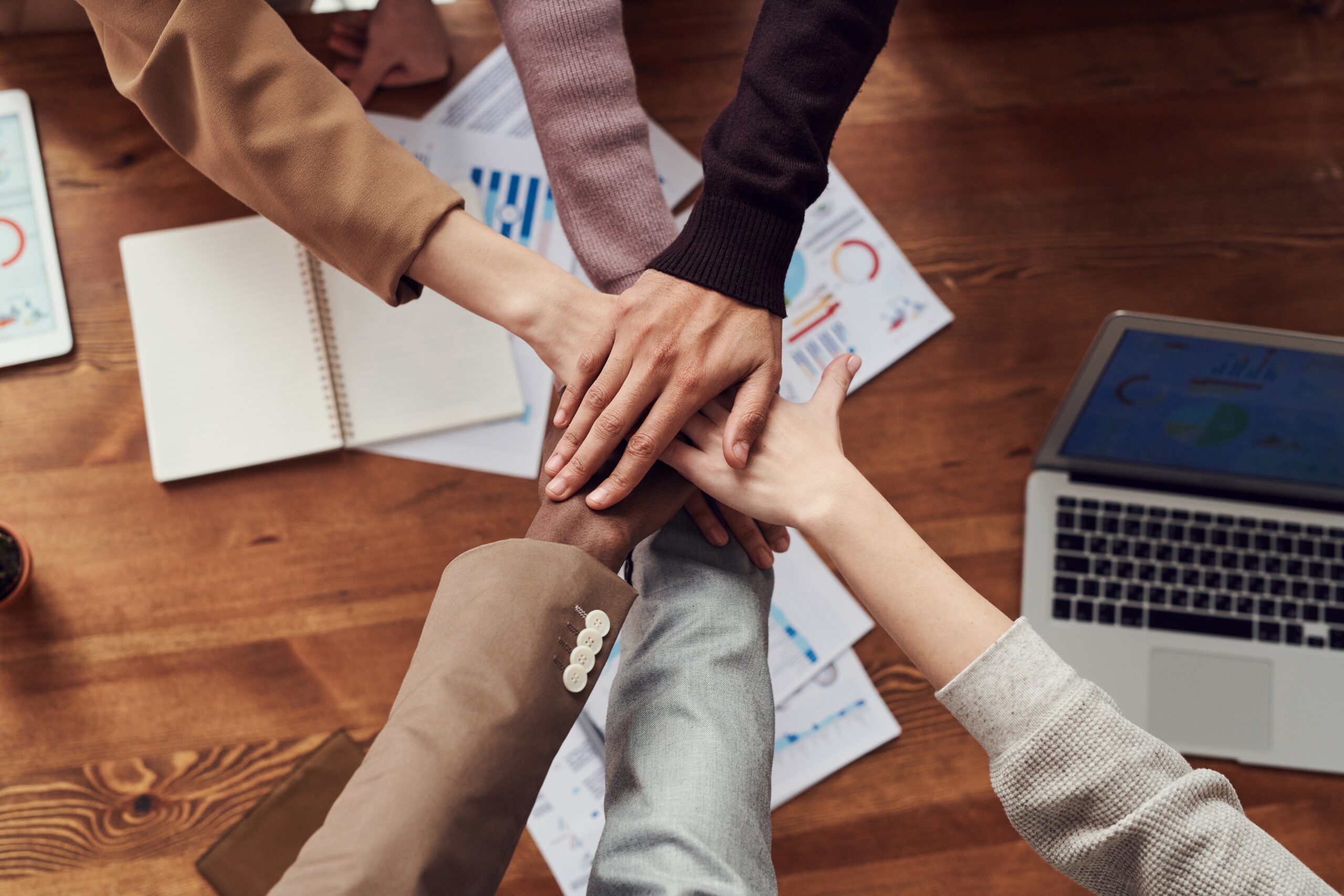 A group of people performing the 'hands in' gesture of teamwork above a desk strewn with papers and a laptop.