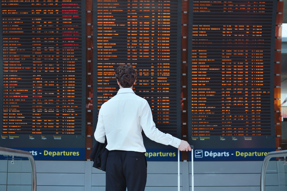 A man in a suit with a travel case looking at a large departures board.